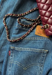 Blaue Jeans aus Denim mit orangefarbener Naht, versehen mit einem roten Levi's-Tag. Eine burgundy-farbene, gepolsterte Handtasche mit einer goldenen Kette ist in der Nähe platziert.