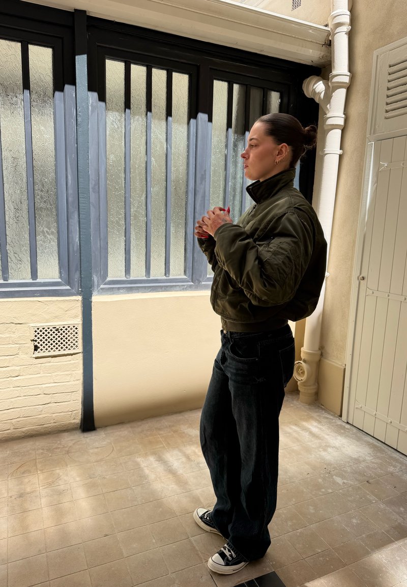 Personne avec les cheveux en chignon portant une veste verte et un pantalon noir, debout à l'intérieur près de fenêtres givrés, les mains jointes.