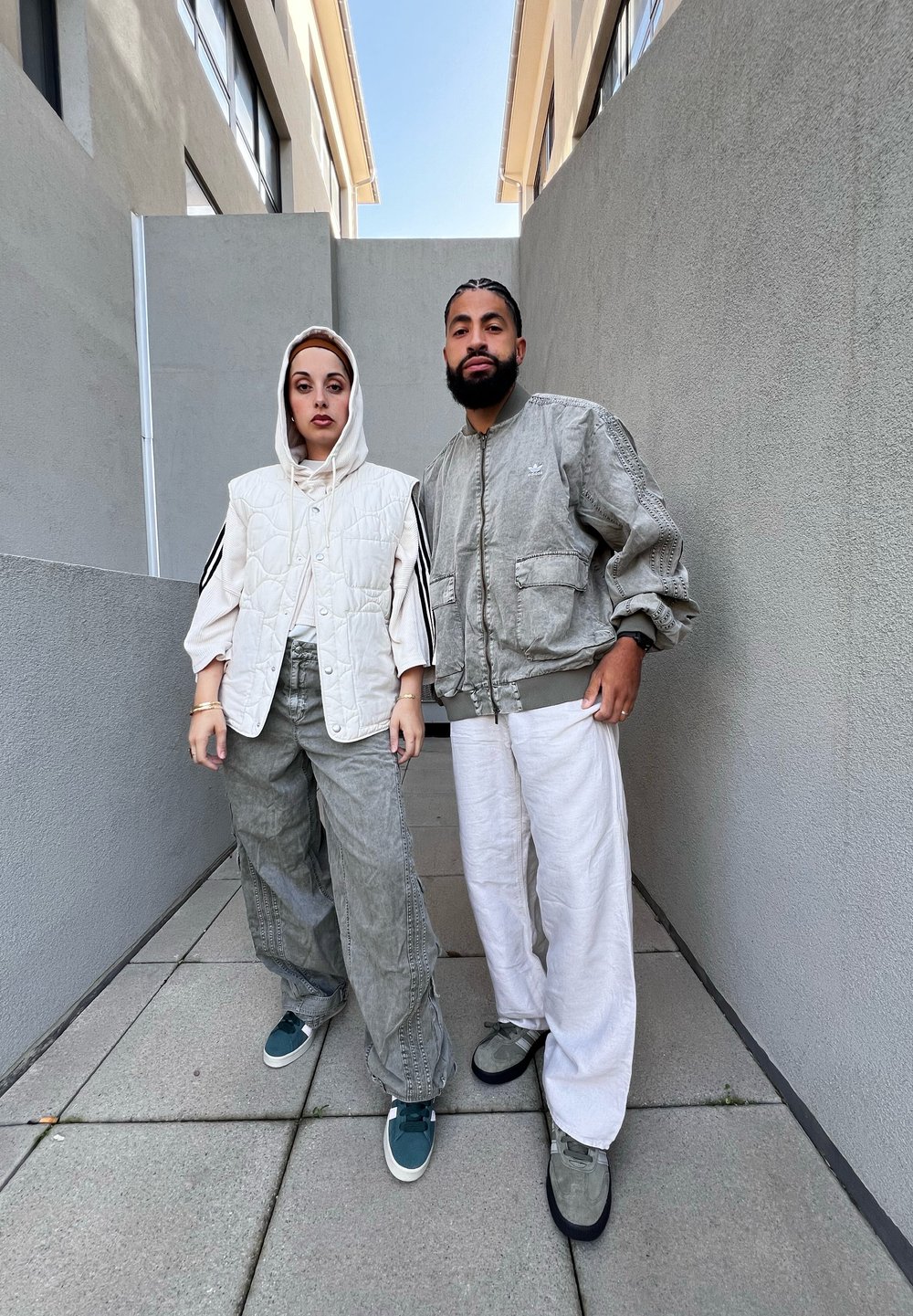 Two people stand on a tiled surface. One wears a white padded vest with a hood, light green baggy pants, and green sneakers. The other wears a gray bomber jacket, white pants, and gray shoes.