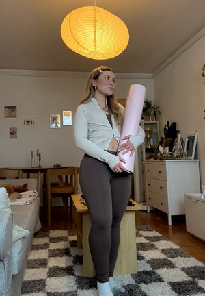 Joven mujer sosteniendo una esterilla de yoga enrollada, de pie en una sala de estar acogedora con una alfombra con estampado y una luz cálida desde arriba.
