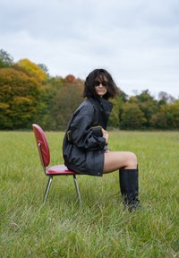 Jachetă neagră supradimensionată cu guler înalt, purtată cu cizme negre până la genunchi. Modelul stă pe un scaun roșu într-un câmp cu iarbă.