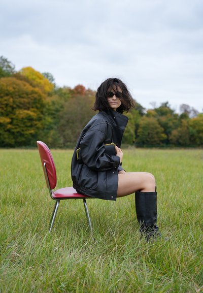 Chaqueta negra de gran tamaño con un cuello alto, combinada con botas negras hasta la rodilla. El modelo se sienta en una silla roja en un campo de hierba.