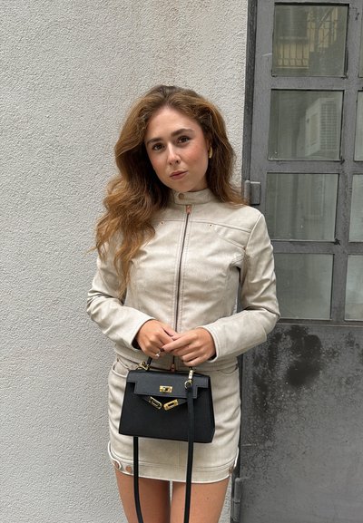 Chaqueta de imitación de cuero beige con un cuello alto y cremallera frontal. Bolso negro con detalles dorados y correa ajustable. Fondo liso.