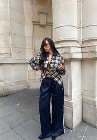 Koszula na guziki w beżowo-czarną kratę na czarnym topie typu bralet; granatowe, szerokie spodnie z satyny z białymi paskami; otoczona kamiennymi kolumnami.