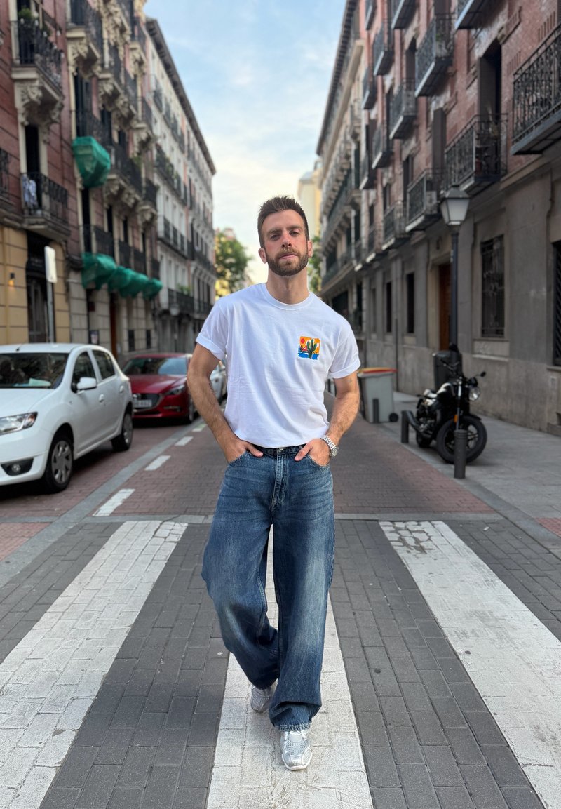 Homme portant un t-shirt blanc et un jean marchant sur un passage pour piétons dans une rue urbaine bordée de voitures garées et de bâtiments.