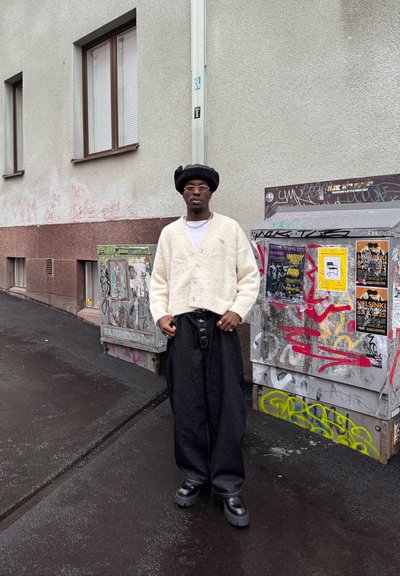 Persona vistiendo un cárdigan beige texturizado y pantalones negros de pierna ancha, complementados con zapatos negros gruesos y un sombrero redondo negro.