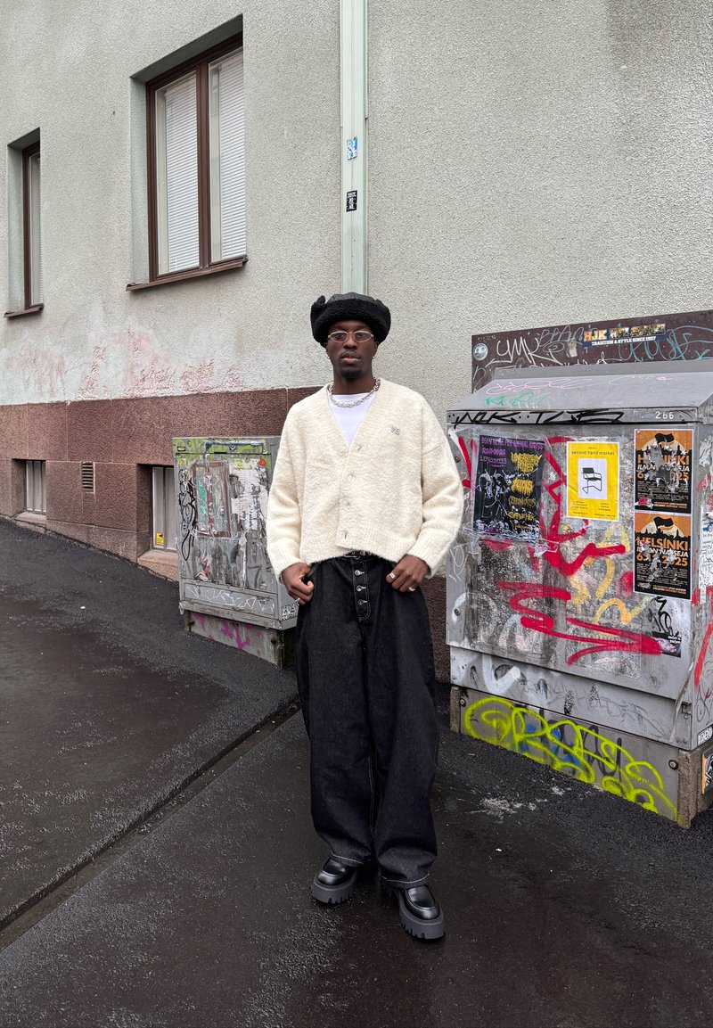 Personne portant un cardigan crème texturé et un pantalon noir à jambes larges, complétés par des chaussures noires épaisses et un chapeau rond noir.