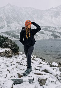 Crna vodootporna jakna, crne tajice i sive planinarske čizme; nosi narančastu i crvenu prugastu kapu; snježni kameniti krajolik i jezero.