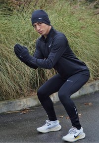 Fekete sportdzseki és leggings fényvisszaverő részletekkel, fekete kötött sapka és kesztyű. Fehér futócipő szürke és sárga részletekkel.