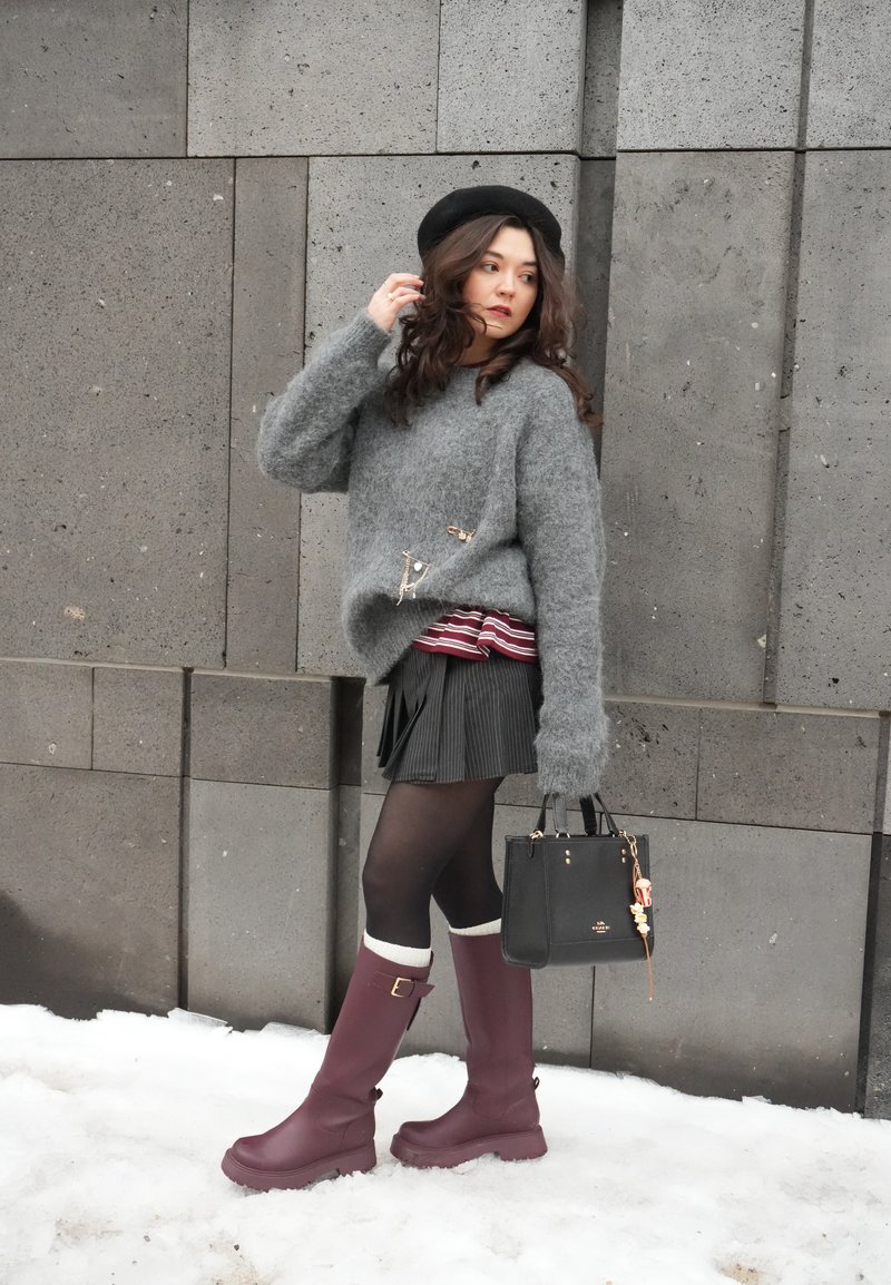 Femme portant un pull gris duveteux, un béret noir, une jupe plissée, des bottes bordeaux, et tenant un sac à main noir, debout sur la neige près d'un mur en pierre.