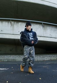 Veste en fausse fourrure grise foncée, hoodie graphique, pantalon camouflage et bottes beiges. Des lunettes de soleil et un bonnet noir complètent le look. Fond en béton.