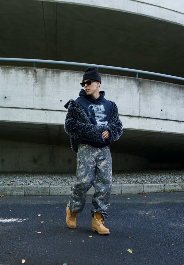 Faux fur jacket in dark gray, graphic hoodie, camouflage pants, and tan boots. Sunglasses and black beanie complete the look. Concrete background.