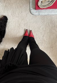 Un pantalon noir ajusté et des talons rouges à bout pointu sont visibles vus d'en haut. L'arrière-plan présente une moquette de couleur claire et une table.