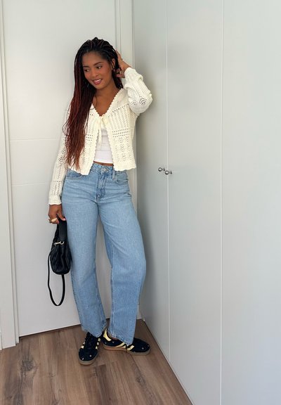 Cardigan de ganchillo en blanco, superpuesto a una blusa blanca. Jeans de pierna ancha en azul claro, zapatos negros con detalles amarillos y un bolso negro.