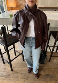 Persona che indossa una giacca di pelle marrone scuro sopra una camicia bianca, jeans azzurri chiari, scarpe marroni, mentre tiene una borsa nera in un ambiente di caffè.