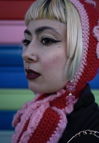 Heklet rød og rosa skjerf, kombinert med matchende lue. Har en nesering og lagdelte øredobber. Fet eyeliner og mørk leppestift.