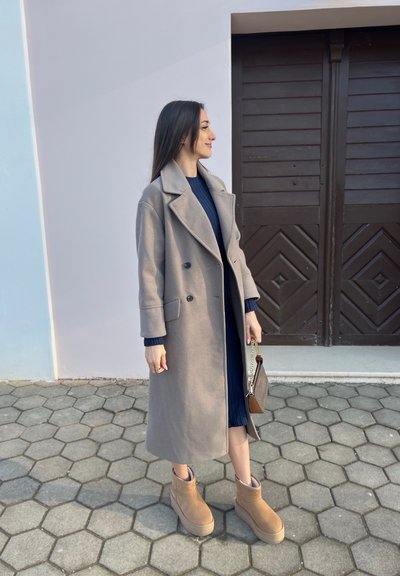 Abrigo gris de lana, de doble botonadura y mangas largas, combinado con un vestido de punto azul marino y botas de tobillo marrones. La modelo lleva un pequeño bolso marrón.