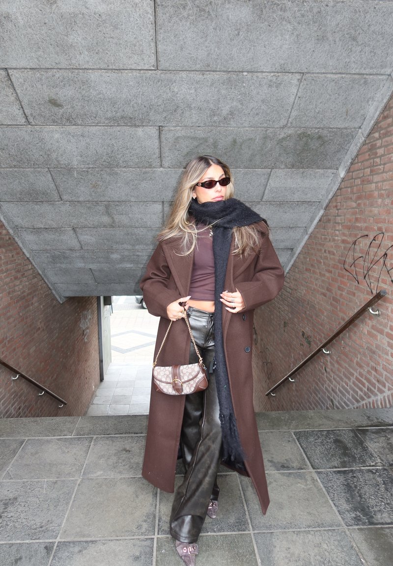 Γυναίκα με μακρύ καφέ παλτό, μαύρο κασκόλ, γυαλιά ηλίου και δερμάτινο παντελόνι, κρατώντας τσάντα με μοτίβο, στέκεται σε περάσμα σκάλας.