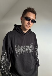 Hoodie noir avec un design de toile d'araignée blanche sur la manche et du texte stylisé en flamme sur la poitrine. Des lunettes de soleil slim et modernes complètent le look.