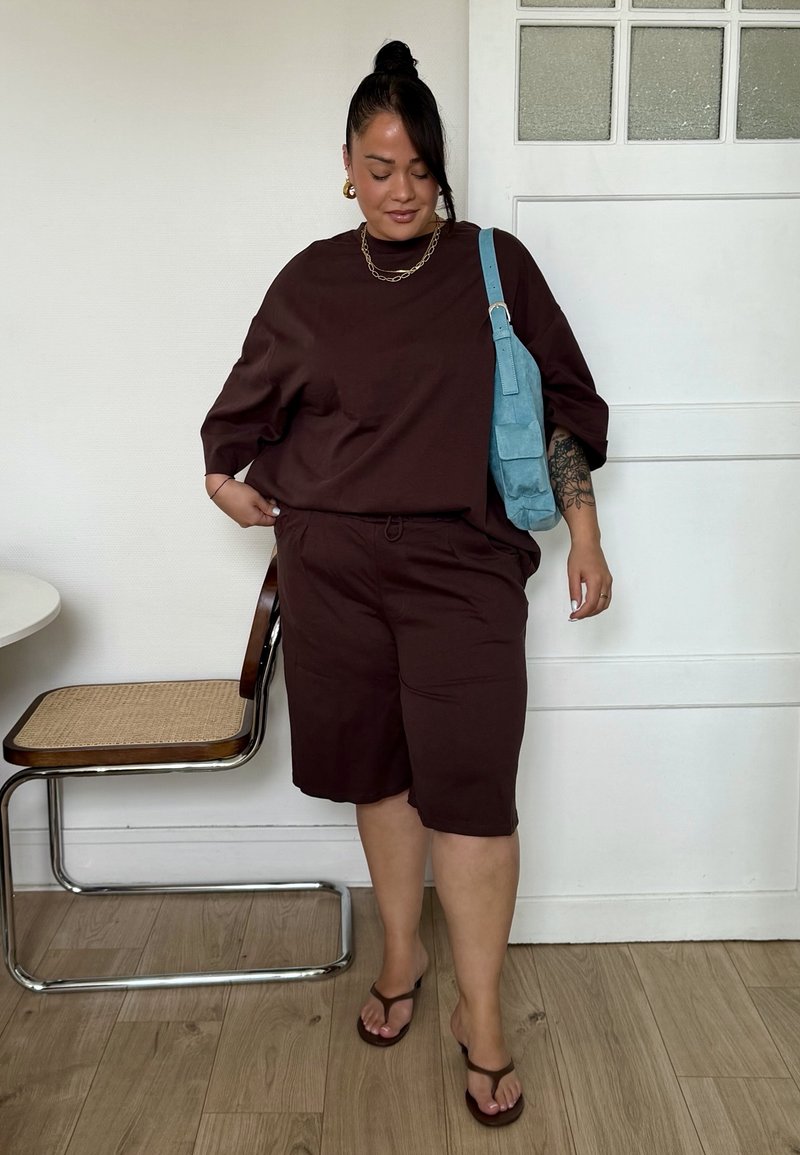 Frau mit dunklen Haaren im Dutt trägt ein lockeres braunes Outfit mit Shorts und Sandalen, trägt eine blaue Schultertasche und steht nahe einem Stuhl und einer Tür.