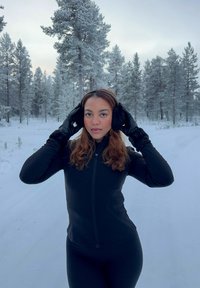 Femme portant une tenue d'hiver noire et des cache-oreilles se tenant sur une route enneigée bordée de pins couverts de neige sous un ciel nuageux.