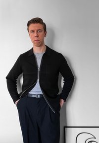 Μαύρος ζιβάγκο με φερμουάρ πάνω από γκρι μπλουζάκι, συνδυασμένος με ναυτικά παντελόνια και μαύρη ζώνη. Απλή ύφανση με λεπτομέρεια τσέπης στο στήθος.