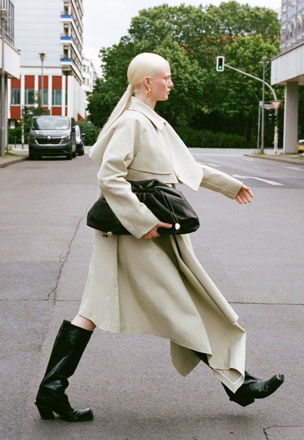 Gai bežs pārmērīgs mētelis ar īsu dizainu, apvienots ar melnu lielu rokassomu un melniem zābakiem līdz ceļiem, staigājot pa ielu.