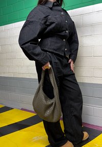 Personne portant une veste en jean noire et un pantalon, tenant un sac à main tissé gris, debout sur un sol à rayures jaunes et noires contre un mur vert et blanc.