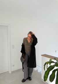 Cappotto nero, top aderente verde oliva, pantaloni grigi a gamba larga, stivali neri massicci, borsa a motivo tenuta in mano. Ambientazione interna luminosa e minimalista.