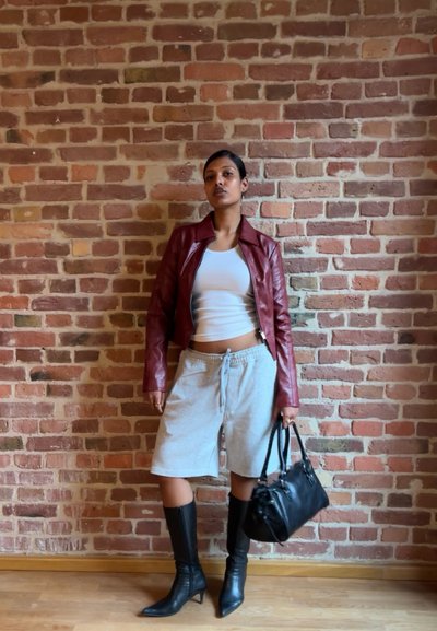 Chaqueta de cuero roja, camiseta blanca ajustada, pantalones cortos grises, botas negras hasta la rodilla y bolso negro. Fondo de pared de ladrillo.