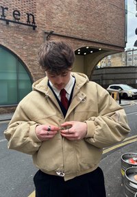 Bézs kapucnis pulóver nagy, fodros ujjakkal és cipzárral az elején. Ezüst kiegészítőkkel és mandzsettákkal díszítve. Egy férfi cipzárt igazít, gyűrűket viselve.