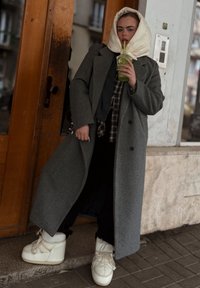 Szürke gyapjúkabát nagy hajtókkal, fekete flanel ingre rétegezve. Fehér platform sportcipő, zöld italt tartva. Fa és kő háttér.