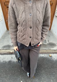 Cardigan marron en maille torsadée avec un col montant et des boutons noirs, superposé à un haut en dentelle noire. Associé à un pantalon large foncé et un sac à main noir.
