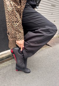 Fekete Nike sportcipő piros talpbetétekkel, laza fekete nadrággal és világosbarna, tigrismintás dzsekivel viselve. Texturált anyag látható.