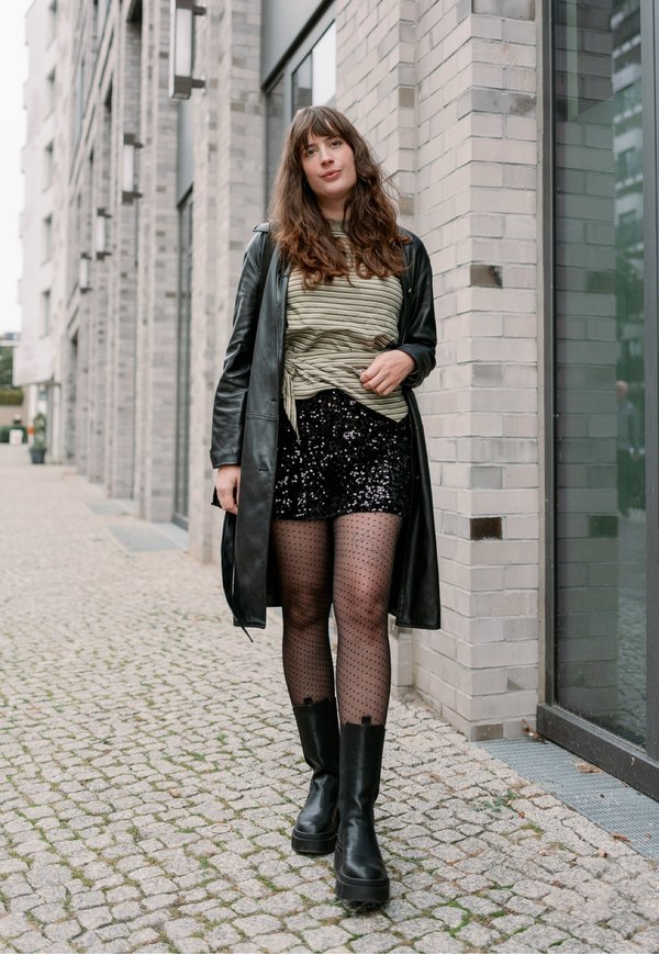 Une femme se tient dans une rue pavée de la ville, près d’un bâtiment en briques. Elle porte un manteau en cuir noir, des collants à motifs et des bottes.