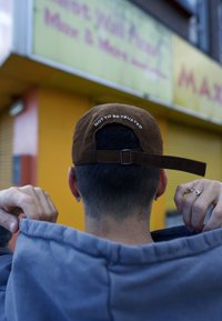 Gorra de pana marrón con el texto blanco "NO CONFIAR," llevada al revés. Sudadera gris, mostrando parte de una mano ajustando el cuello.