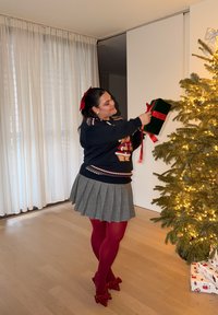 Una donna con un maglione scuro con un disegno di orso e una gonna grigia plissettata tiene una scatola regalo verde con un nastro rosso vicino a un albero di Natale.