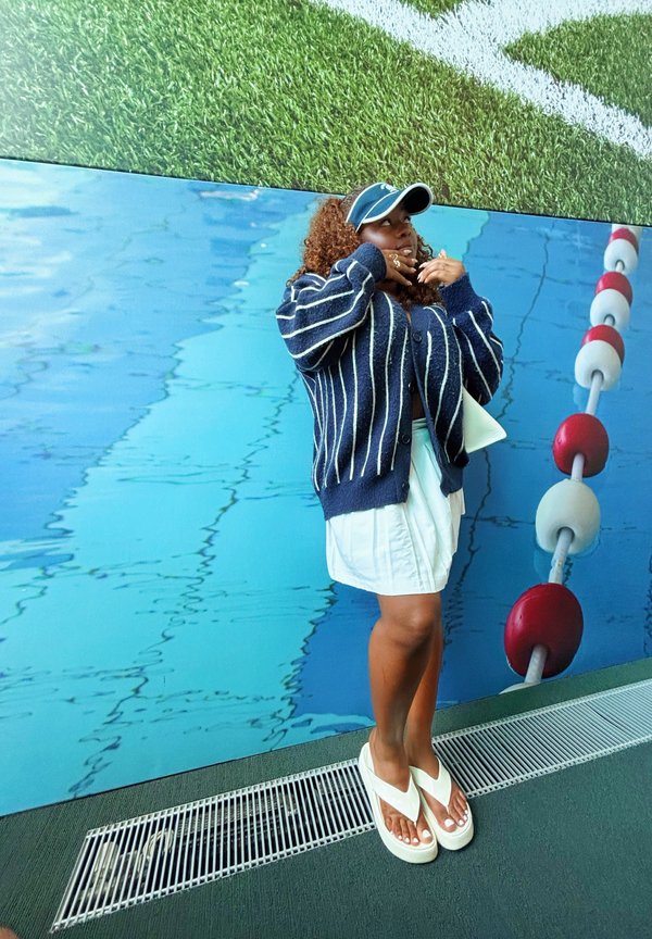 Marine gestreifter Cardigan, weißes plissiertes Kleid und weiße Flip-Flops. Der Hintergrund zeigt einen blauen Swimmingpool mit roten und weißen Bahnmarkierungen.