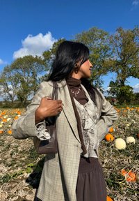 Plaid grey blazer over a brown ruffled top, paired with a brown skirt. Holding a dark brown handbag. Field of pumpkins in the background.