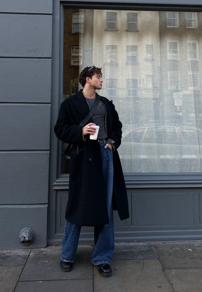 Abrigo de lana negro, suéter gris y pantalones de mezclilla de pierna ancha. Sosteniendo una taza de café blanca, de pie junto a una ventana con un fondo de pared gris.