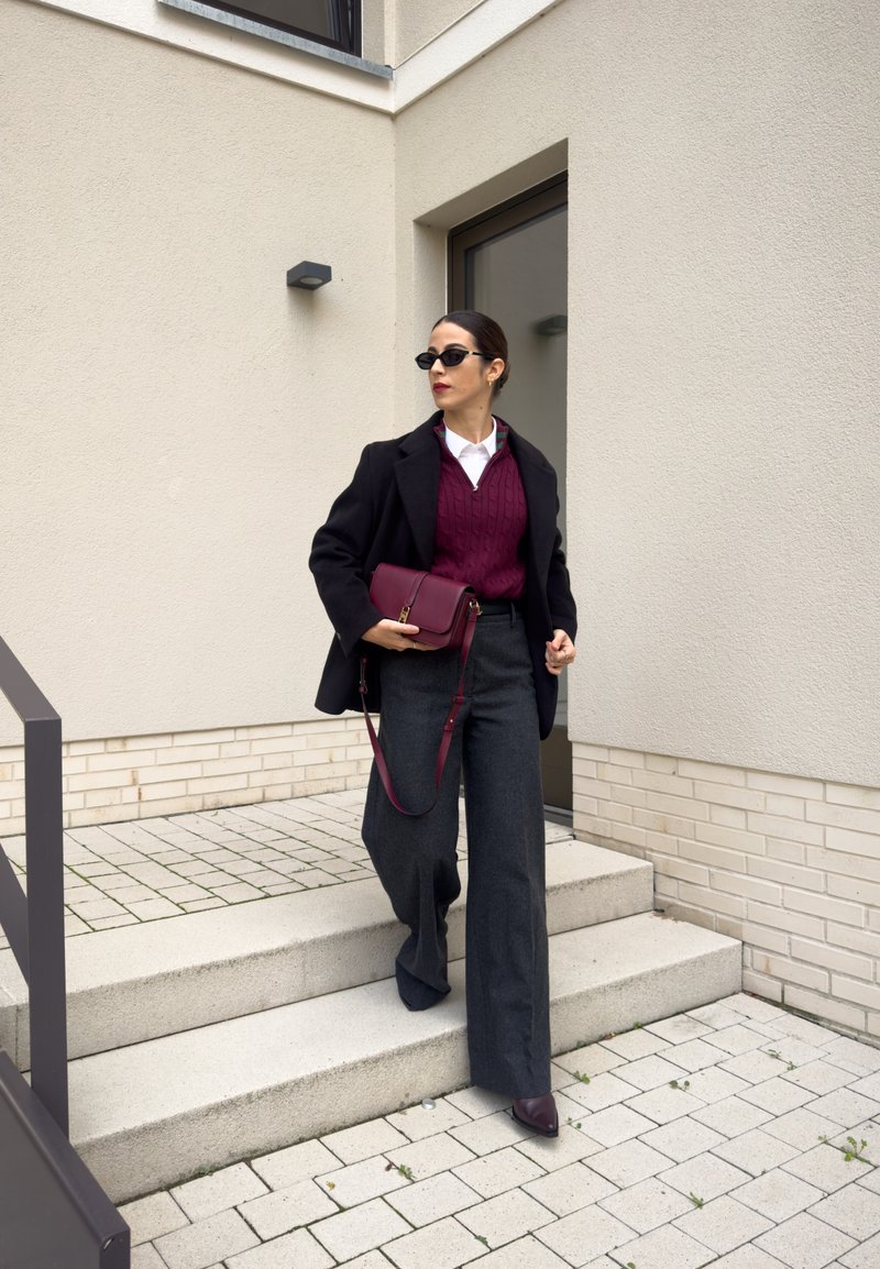 Hallitusmaterjalist hallid laiad püksid, burgundia värvi palmikutega sviiter, must kahepoolselt nööpidega mantel ja marooni värvi õlakott, mida kaunistavad prillid.