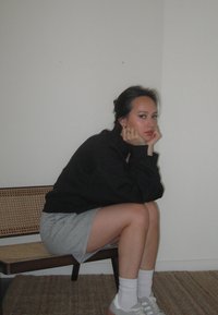 Μαύρη μπλούζα, γκρι φούστα, λευκές κάλτσες και αθλητικά παπούτσια. Καθισμένος σε μια ξύλινη καρέκλα με πλέκη πλάτη. Απλή, μινιμα λιστική διακόσμηση.