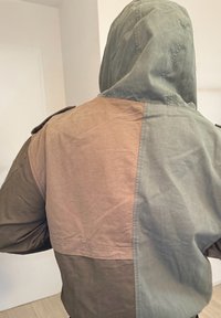 Personne portant une veste à capuche avec des blocs de couleur verts, marron et beige, debout à l'intérieur, dos à la caméra.