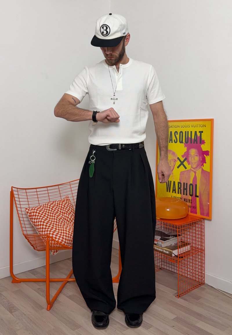 Hombre con gorra blanca y camisa con collar de cruz mira su reloj, de pie junto a una silla de alambre naranja con cojín a cuadros y póster de arte colorido.