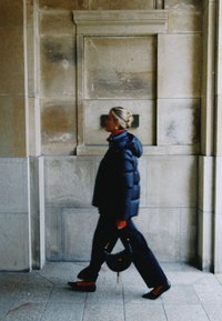 Kvinna som går förbi en stenmur, klädd i en svart vadderad jacka, svarta byxor, röda strumpor, svarta skor och bär en svart handväska.