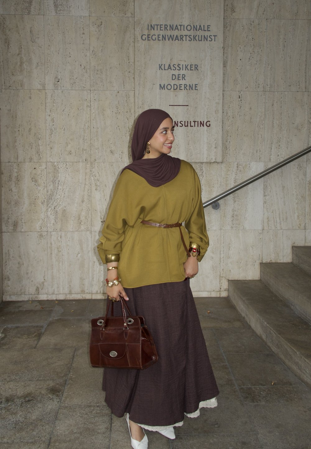 Olivově zelený oversized top s zlatými manžetami a hnědým páskem, spárovaný s tmavě hnědou dlouhou sukní a bílými zdobenými botami. Hnědá kabelka.