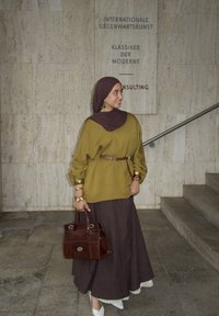 Olivově zelený oversized top s zlatými manžetami a hnědým páskem, spárovaný s tmavě hnědou dlouhou sukní a bílými zdobenými botami. Hnědá kabelka.