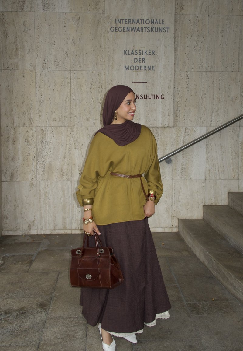 Blusa grande de color verde oliva con puños dorados y cinturón marrón, combinada con una falda larga marrón oscuro y zapatos blancos decorados. Bolso marrón.