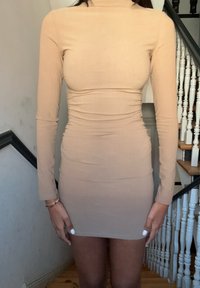 Vestido justo, de manga longa, feito de tecido elástico em bege. Apresenta um decote alto e detalhes franzidos ao longo dos lados. Textura suave.