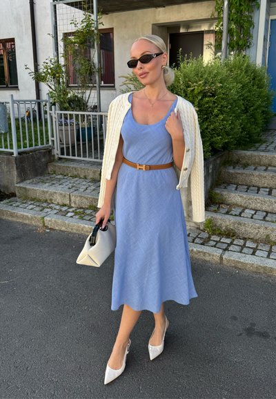 Vestido sin mangas azul claro con un patrón texturizado, ceñido a la cintura con un cinturón marrón. Combinado con un cárdigan crema y tacones blancos.